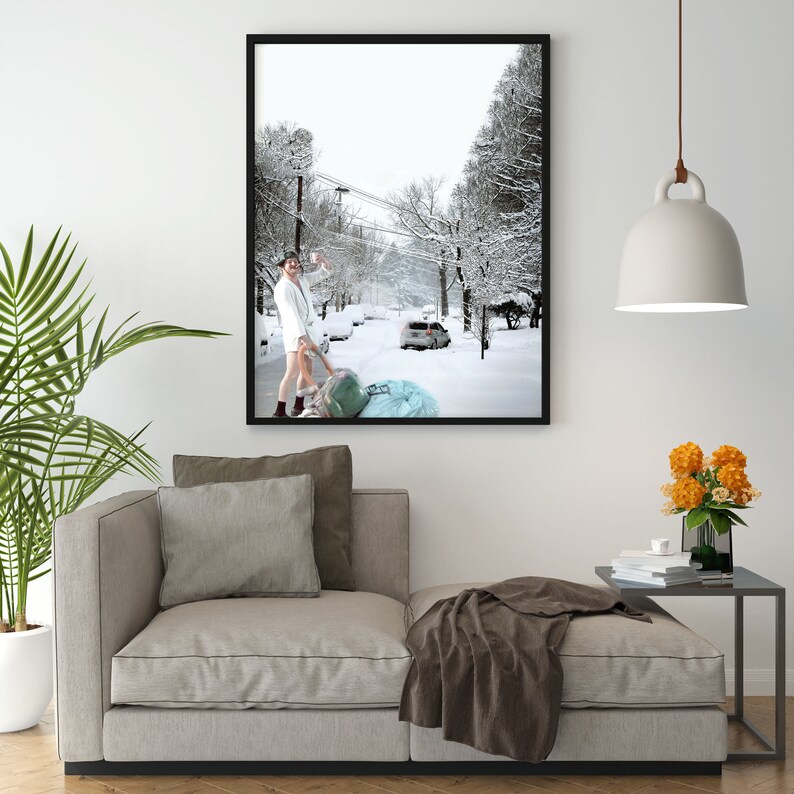 May include: A man in a white robe walks down a snowy street, pulling a large blue bag behind him. The street is lined with trees and houses, and the snow is falling softly.