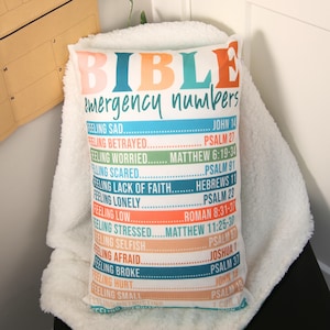 Könnte beinhalten: Ein weißes Kissen mit den Worten "BIBLE emergency numbers" in bunten Buchstaben bedruckt. Das Kissen zeigt eine Liste von Bibelversen für verschiedene Gefühle, wie z. B. Traurigkeit, Verrat, Besorgnis, Angst, Mangel an Glauben, Einsamkeit, Niedergeschlagenheit, Stress, Egoismus, Angst, Armut, Verletzung, Kleinheit und Misstrauen.