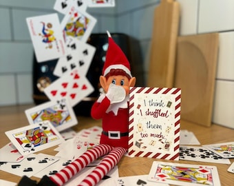 Può includere: Una bambola elfo rossa e bianca con un cappello a punta, seduta tra carte da gioco sparse. L'elfo tiene in mano un cartoncino bianco con bordi a strisce rosse con la scritta, I think I shuffled them too much, sotto una cascata di carte.