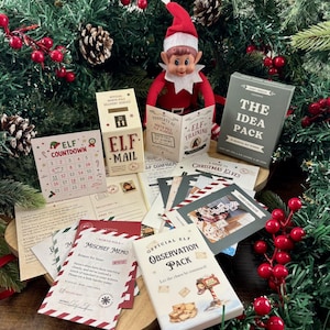 Pu&ograve; includere: Una bambola elfo di Natale con cappello e tuta rossa siede dietro una collezione di articoli di carta stampata, tra cui una scatola Elf Mail, un calendario Elf Countdown, un Mischief Memo, un Observation Pack e The Idea Pack con varie carte.