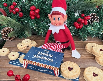 Può includere: Una bambola elfo rossa e bianca con un cappello rosso e gambe a righe è seduta su una superficie di legno circondata da biscotti e verde. Una scatola blu con la scritta "The Magical Maximizer" è aperta.