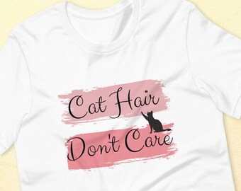 Pode incluir: Camiseta branca com um design de pinceladas rosa e o texto "Cat Hair Don't Care" em preto. Uma silhueta preta de um gato está incluída no design.