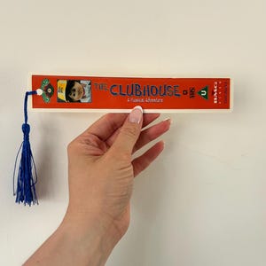 May include: A rectangular bookmark with the text "THE CLUBHOUSE A Musical Adventure" and a child's picture. The bookmark has a red and white design and a blue tassel attached.
