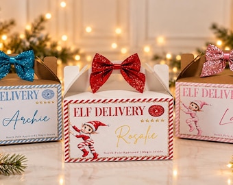 May include: Three rectangular cardboard gift boxes labeled ELF DELIVERY with five stars and a North Pole stamp. Each box features a cartoon elf, a personalized name in script, and a glittery bow on top in blue, red, or pink.