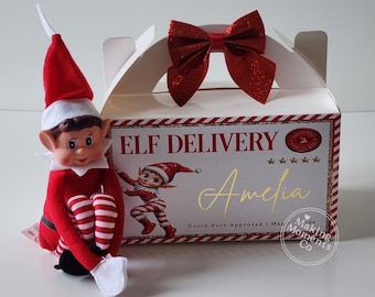May include: A plush elf doll with a red hat and striped legs sits beside a white cardboard gift box. The box features a red glittery bow, the text "ELF DELIVERY", "Amelia", "North Pole Approved", and a graphic of an elf.