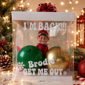 May include: A clear square gift box containing a small elf doll wearing a red hat and suit, positioned between a large green ornament and a large gold ornament. The box features white text reading I'M BACK!!!, Brodie, and GET ME OUT.