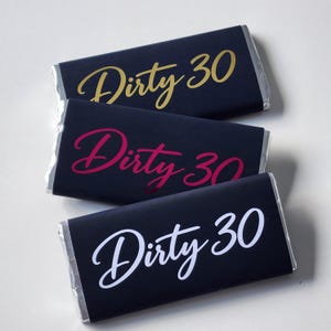 May include: Three rectangular chocolate bars wrapped in black paper with the text Dirty 30 printed in a cursive font. The top bar features gold text, the middle bar features magenta text, and the bottom bar features white text on the black wrapper.