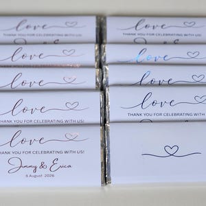 May include: Ten rectangular chocolate bars wrapped in white paper with metallic foil accents. The wrappers feature the word love in cursive script, a small heart icon, and the text THANK YOU FOR CELEBRATING WITH US. One wrapper includes the names Jimmy & Erica and the date 8 August 2026.