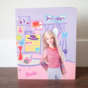 May include: A Barbie-themed binder with a cartoon illustration of a dressing room. The cover features a Barbie doll in a striped shirt and jeans. The background is lavender with pink accents and the word "Dressing Room".