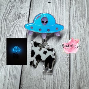 May include: A badge reel featuring a blue and purple alien spaceship design with a purple alien head. Below the spaceship is a black and white cow. The badge reel has a clear plastic clip.