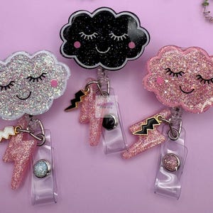 May include: Three badge reels featuring cloud-shaped tops with glittery finishes and smiling faces with closed eyes. Each cloud has a dangling lightning bolt charm attached. The clouds are silver, black, and pink, with clear plastic straps.