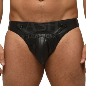 Puede incluir: Jockstrap de cuero negro con una bolsa contorneada y detalles de broches. El jockstrap presenta el texto "ASH LEATHERS" y está diseñado para un ajuste ceñido. La prenda está hecha de cuero liso y oscuro.