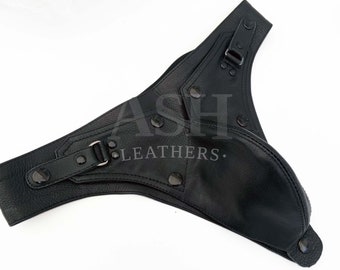 May include: A black leather codpiece featuring a contoured pouch design with adjustable straps, metal buckles, and dark hardware studs. The surface is embossed with the text ASH LEATHERS across the center, showcasing a textured finish.
