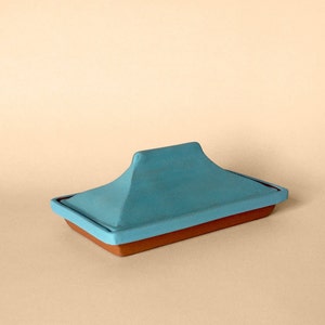 May include: A blue ceramic butter dish with a lid. The dish is rectangular and has a raised edge. The lid is shaped like a dome and has a pointed top.