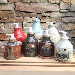 May include: A collection of seven ceramic soap dispensers with white plastic pumps. The dispensers feature various finishes including solid red, speckled cream, light blue, and brown glazes, with some displaying detailed mountain and camping illustrations.