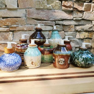 May include: A collection of nine ceramic soap dispensers with various glaze finishes, including blue speckled, brown, turquoise, and green patterns. Each dispenser features a white plastic pump mechanism and a cork stopper, arranged on a wooden surface.