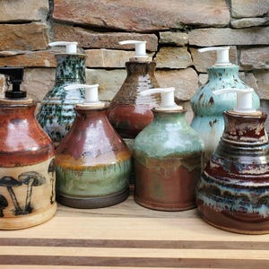May include: A collection of six ceramic soap dispensers with various glaze finishes, including a cream-colored vessel with black mushroom illustrations, solid earth tones, and mottled blue-green patterns, each topped with a functional pump mechanism.