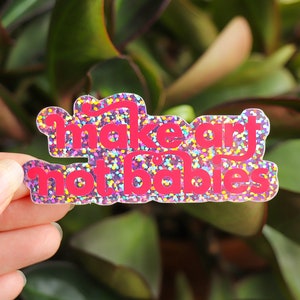 Könnte beinhalten: Ein rosa Glitzeraufkleber mit dem Text "Make art not babies".