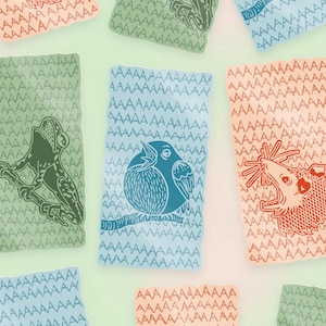 Op de afbeelding: Een set van negen kleurrijke kaarten met een herhalend patroon van de letter 'A'. Elke kaart heeft een andere dierenillustratie: een kikker, een vogel en een opossum. De kaarten zijn gedrukt op een getextureerd papier.