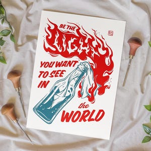Peut inclure: Une impression d'art rectangulaire blanche représentant une bouteille en verre bleu avec une mèche en tissu, entourée de flammes rouges. Le texte indique : BE THE LIGHT YOU WANT TO SEE IN THE WORLD. Un petit logo carré figure en haut à droite.