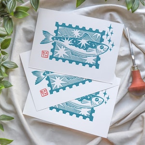 May include: Three white cards with teal fish illustrations resembling postage stamps. Each fish design includes stars and a small red square with text. A carving tool with a wooden handle and a sprig of green leaves are also visible.