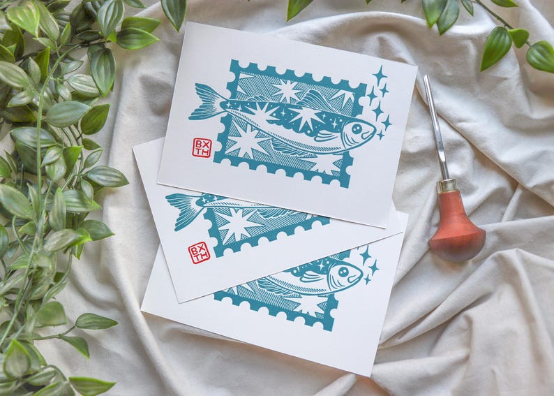 May include: Three white cards featuring teal fish illustrations in a postage stamp design. Each fish design includes stars and a small red square with text. A carving tool with a wooden handle and green leaves are also visible.