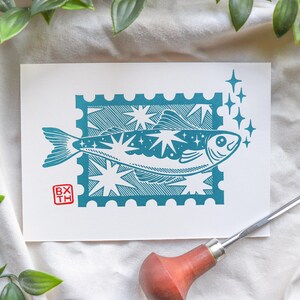 May include: A teal and white art print with a fish design inside a postage stamp border. The fish is decorated with stars and surrounded by sparkling accents. The print includes the initials "BX TH" in a red square.