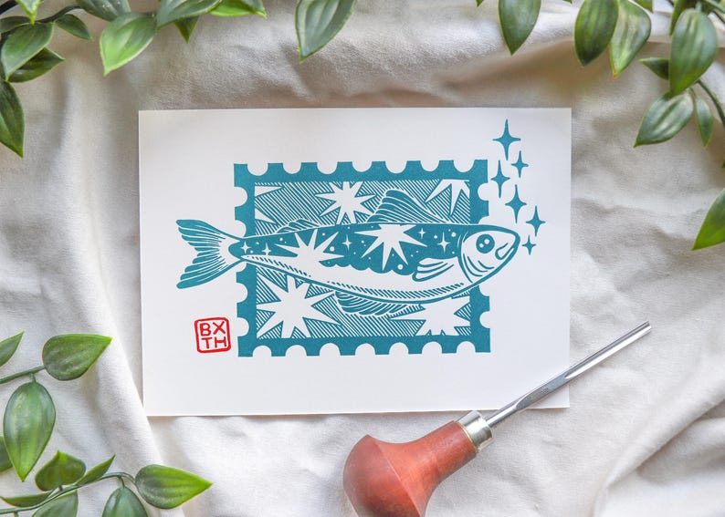 May include: A teal and white art print with a fish design inside a postage stamp border. The fish is decorated with stars and surrounded by sparkling accents. The print includes the initials "BX TH" in a red square.
