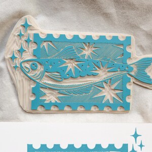 May include: A turquoise fish design wood stamp with starbursts and a postage stamp border. The stamp is on a white surface. The fish is stylized and the design is carved into the wood. The stamp is a unique art supply.
