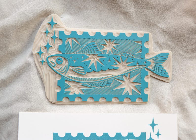 May include: A turquoise fish design wood stamp with starbursts and a postage stamp border. The stamp is on a white surface. The fish is stylized and the design is carved into the wood. The stamp is a unique art supply.