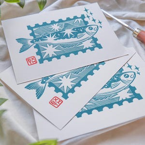 May include: Three white art prints with a teal fish design in a postage stamp frame. The fish is surrounded by stars. Each print has a red stamp with the letters "BX TH". A carving tool with a wooden handle is in the background.
