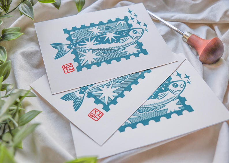 May include: Three white art prints with a teal fish design in a postage stamp frame. The fish is surrounded by stars. Each print has a red stamp with the letters "BX TH". A carving tool with a wooden handle is in the background.