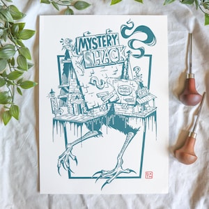 May include: A teal and white print of the Mystery Shack, featuring the text "Mystery Shack" and "World Famous Gifts." The artwork depicts the building with bird-like legs and claws. Two wood carving tools are visible.