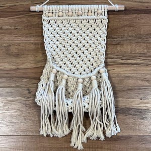 May include: A cream-colored macrame wall hanging featuring a diamond-patterned weave, suspended from a light-toned wooden dowel with a cord. The piece is accented with natural wooden beads along the top and bottom, finished with five long tassels.