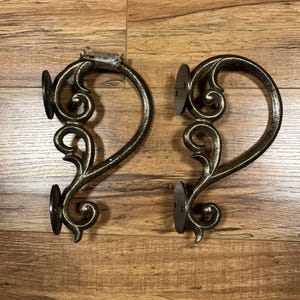 May include: Two matching metal wall brackets with a dark bronze finish and a decorative scrollwork design. Each bracket features two circular mounting plates with pre-drilled holes for installation, showcasing a curved, ornate, and symmetrical shape.