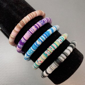 May include: Five colourful bracelets made of stacked round beads. The bracelets feature different geometric patterns in shades of pink, blue, white, black, and grey.