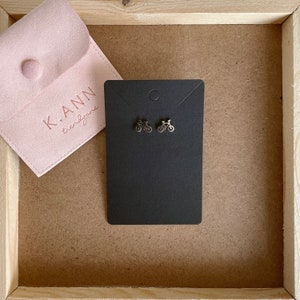 Puede incluir: Un par de pendientes de bicicleta plateados en una tarjeta negra con el texto "K.ANN trendzone" en una bolsa de gamuza rosa.