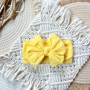 May include: A bright yellow baby headband with a large bow. The headband is made of a textured fabric and is centered on a white macrame placemat with fringe. The background includes other macrame elements in neutral tones.