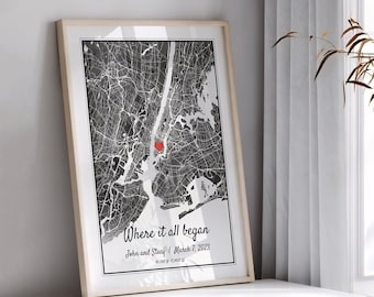 May include: A framed vertical map print featuring a black and white street grid of New York City with a small red heart icon. The text reads Where it all began, John and Stacy, March 7, 2023, and coordinates. The print is in a light wood frame.