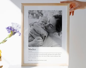 May include: A framed print featuring a black and white photograph of an adult holding a baby. Below the image, the text reads: Mother [Noun] A person who loves unconditionally and puts the needs of her children before her own. See also: caring, superwoman, saint.