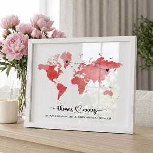 Può includere: Una stampa incorniciata in bianco che mostra una mappa del mondo ad acquerello rosso con una linea tratteggiata che collega due cuori neri. Sotto la mappa, i nomi Thomas e Nansy sono scritti in corsivo con un cuore, seguiti dal testo: DISTANCE MEANS SO LITTLE, WHEN YOU MEAN SO MUCH.