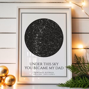 Op de afbeelding: Een ingelijste print met een zwart-witte sterrenkaart. De tekst "UNDER THIS SKY YOU BECAME MY DAD" staat eronder, met details over locatie en datum. Gouden bollen en lichtslingers staan op de voorgrond. De lijst is wit.