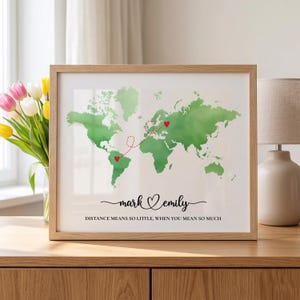 Op de afbeelding: Een ingelijste print met een groene aquarel wereldkaart met twee rode harten verbonden door een rode stippellijn. Onder de kaart staan de namen mark en emily in zwarte schrijfletters, gevolgd door de tekst DISTANCE MEANS SO LITTLE, WHEN YOU MEAN SO MUCH.