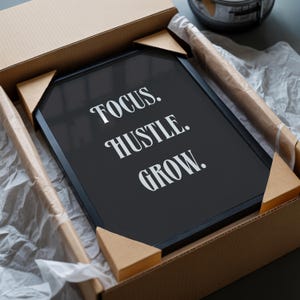 Pu&ograve; includere: Un quadro rettangolare con cornice nera e testo bianco che recita FOCUS. HUSTLE. GROW. La stampa incorniciata &egrave; imballata in modo sicuro all'interno di una scatola di spedizione in cartone marrone con angoli protettivi e carta velina.
