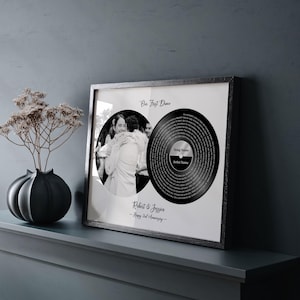 May include: A framed black and white print featuring a photo and a vinyl record design. The photo shows a couple embracing. The vinyl record design includes the text "Our First Dance", "Song Name", and "Artist Name". The frame also includes the names "Robert & Jessica" and "Happy 2nd Anniversary".