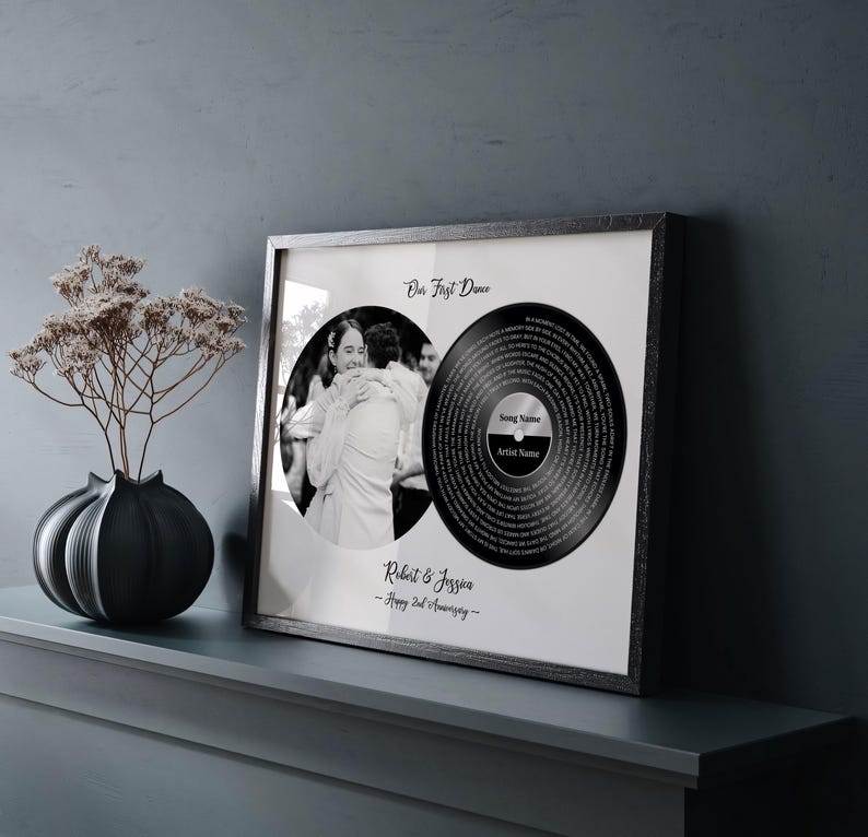 May include: A framed black and white print featuring a photo and a vinyl record design. The photo shows a couple embracing. The vinyl record design includes the text "Our First Dance", "Song Name", and "Artist Name". The frame also includes the names "Robert & Jessica" and "Happy 2nd Anniversary".