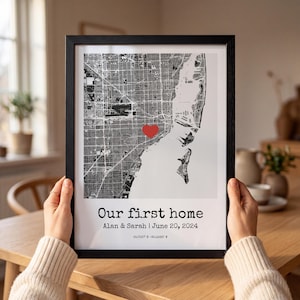 May include: A framed map print featuring a black and white street grid with a red heart icon. The text reads Our first home, Alan & Sarah | June 20, 2024, and 25.7257 N -80.2265 W. The print is enclosed in a thin black frame with a white border.