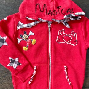 May include: Red zippered hoodie with black and white checkered fabric accents, Mickey Mouse applique, and embroidered text "Magical" on the hood.