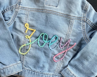 Könnte beinhalten: Eine hellblaue Jeansjacke mit dem Namen "Joey", der mit regenbogenfarbenem Garn auf der Rückseite gestickt ist.