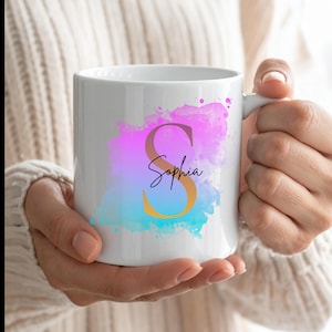 Könnte beinhalten: Ein weißer Keramikbecher mit einem farbenfrohen Aquarell-Spritzer-Design in Rosa-, Lila- und Blautönen. In der Mitte des Spritzers befindet sich ein großer goldfarbener Buchstabe S, über den der Name Sophia in schwarzer Schreibschrift gedruckt ist.
